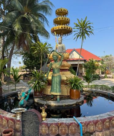 A colorful temple courtyard with statues, tropical plants, and red-roofed buildings